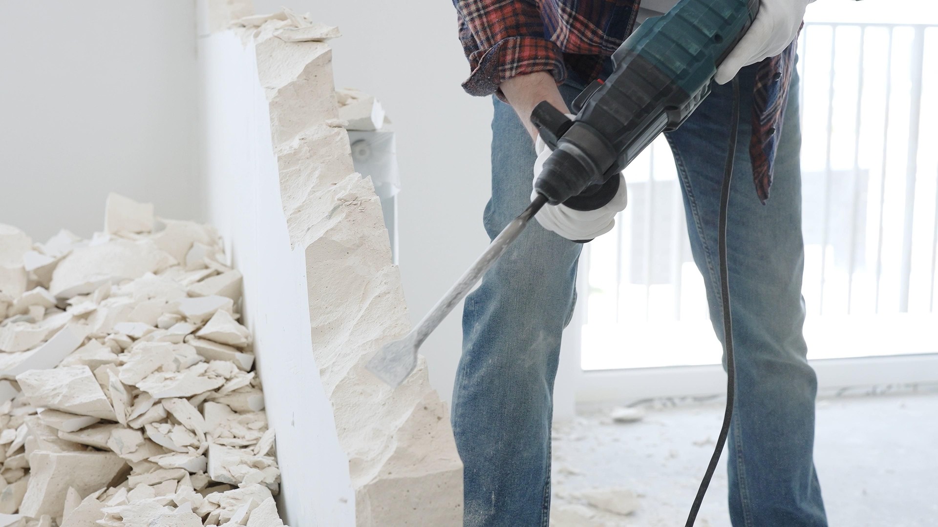 Nahaufnahme eines unbekannten männlichen Bauarbeiters, der rotkarierte Hemdjeans und Schutzhandschuhe trägt, reißt mit einem Bohrhammer eine weiße Wand ab und erzeugt Trümmer auf dem Boden. Renovierung
